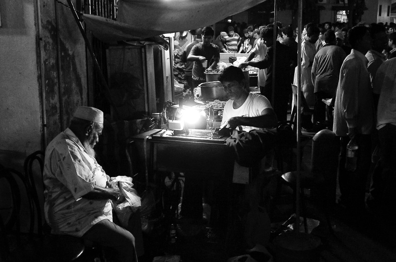A tailor is stitching a cloth in his sewing machine at night with another man assisting him. Bangla Bazar, Singapore