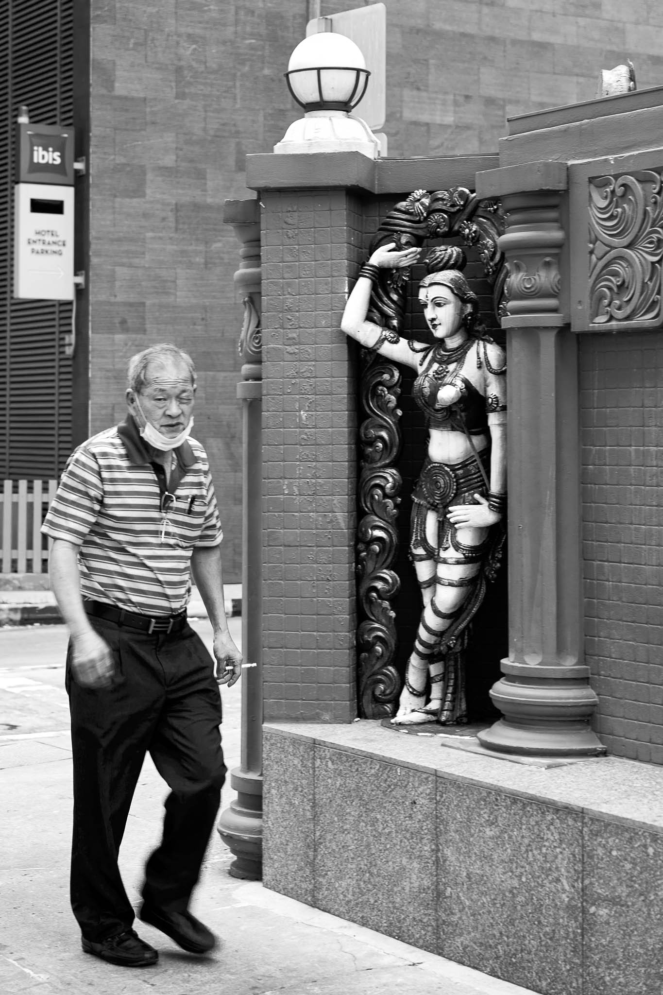 An old pedestrian walking past  the rear gate of the Sri Krishnan Temple, watched over by a guardian deity statue, Bugis, Singapore
